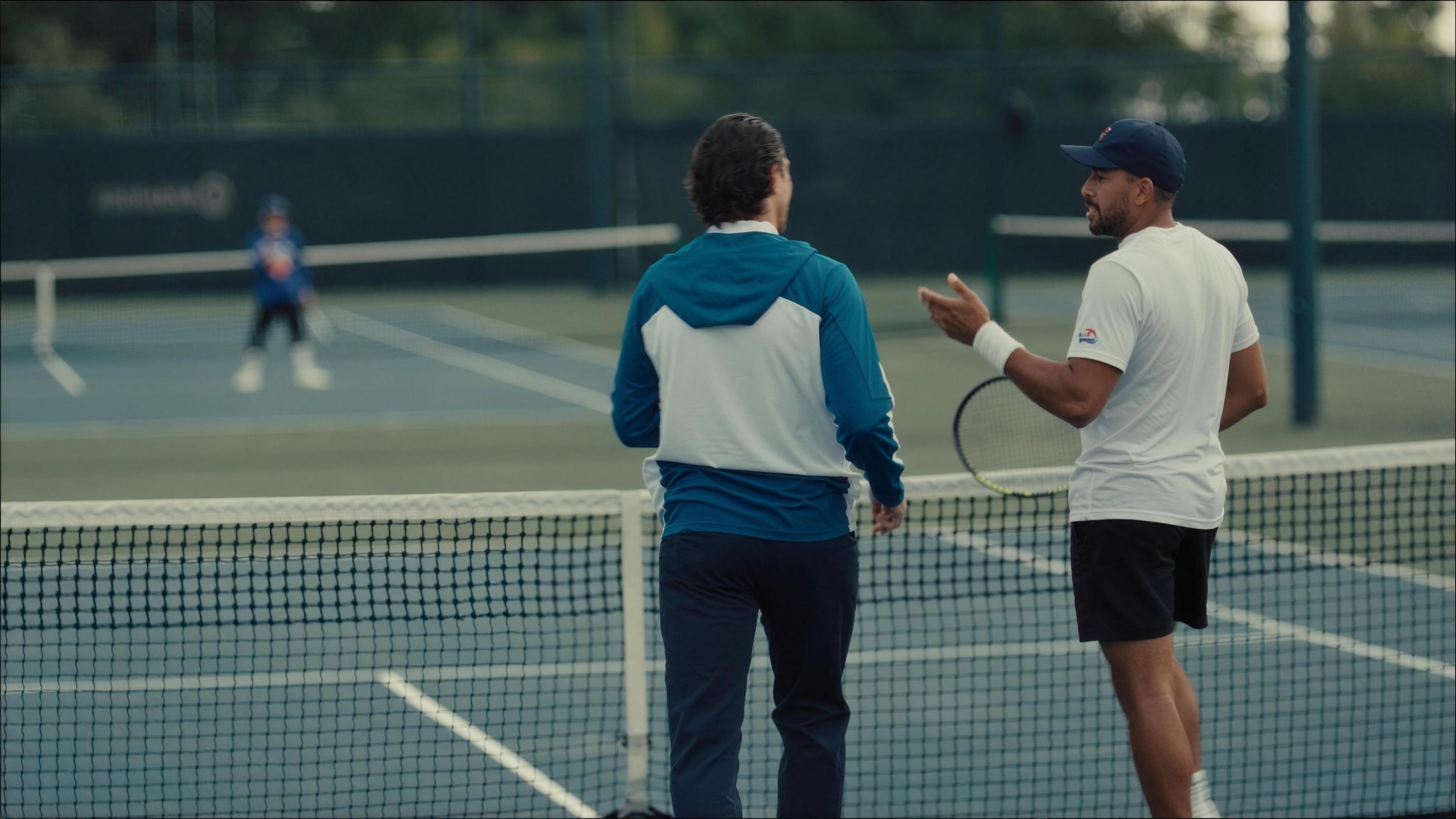 Two tennis professionals talking at the net, a player training beyond
